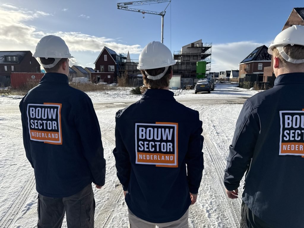 Drie vakmensen met witte helm en werkjas van Bouwsector Nederland, van achteren, op een besneeuwde nieuwbouwplaats